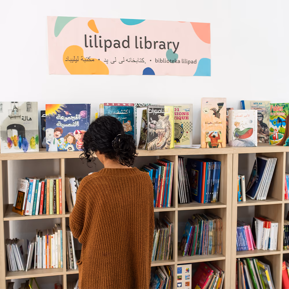 Person in a brown sweater browsing books on shelves under a Lilipad Library banner in a children's library.