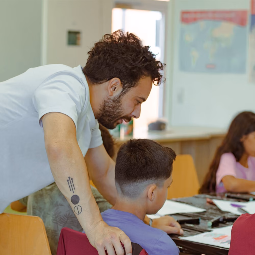 Teacher with tattoos leaning over to help a student in a classroom with other children working on art projects.