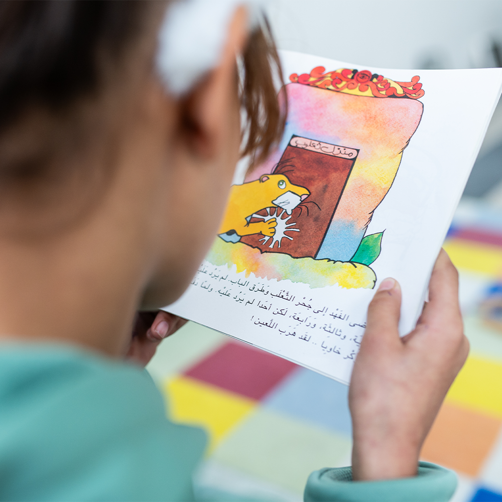 Un enfant en train de lire un livre illustré comportant un texte en arabe et le dessin d'un animal jaune tenant un objet blanc près d'une porte colorée.