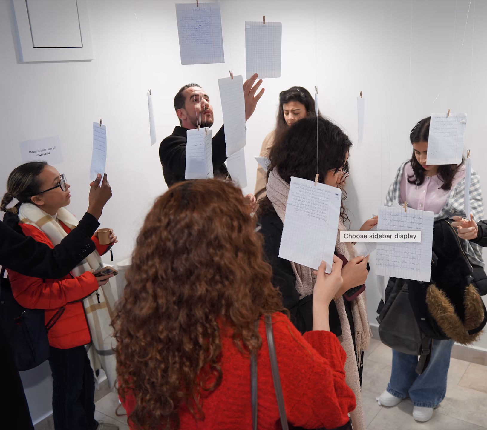 Group of people holding up colorful childlike drawings of faces in front of their own faces indoors.