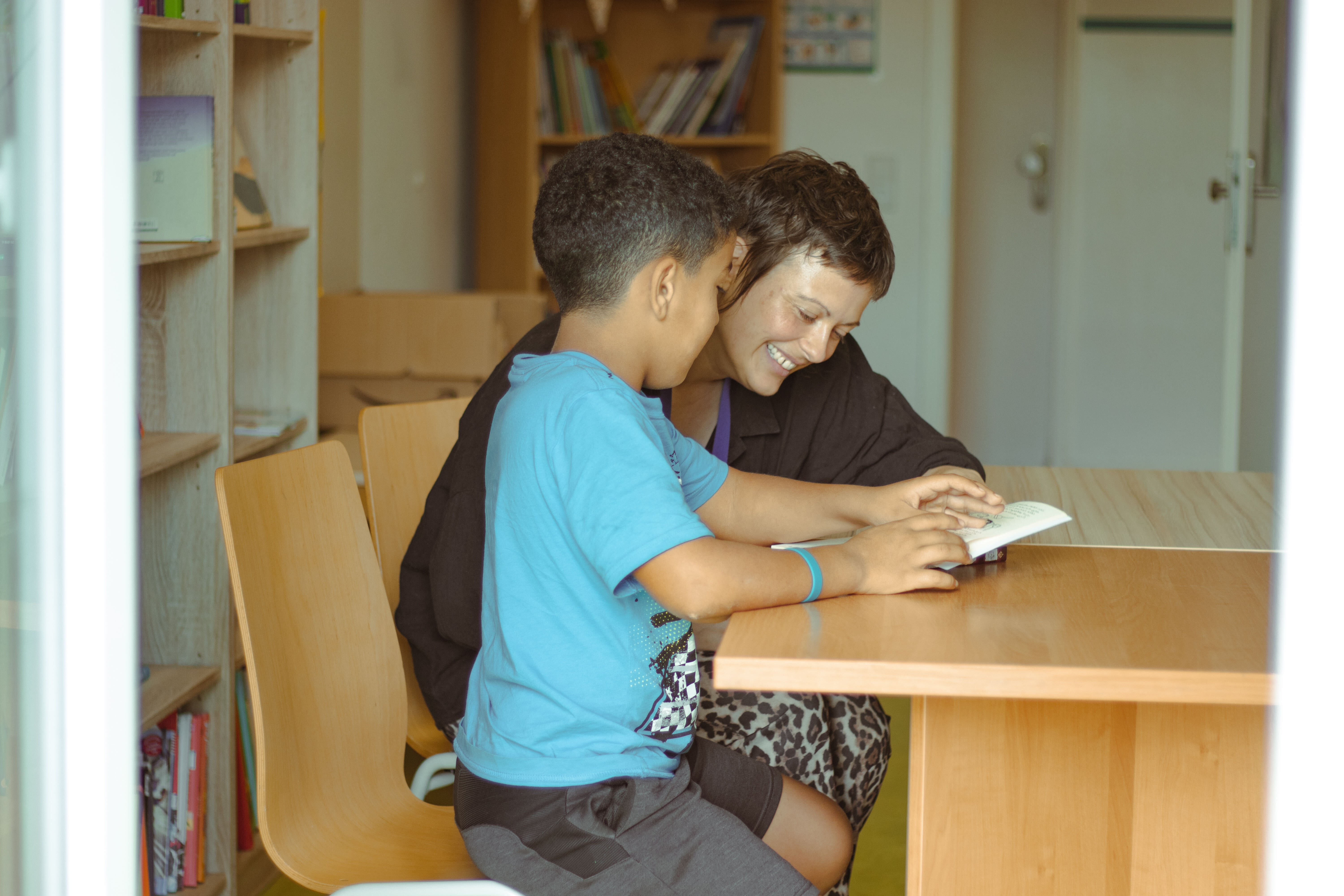 Three children seated in a library holding colorful books in front of their faces, with bookshelves and stacks of books behind them.