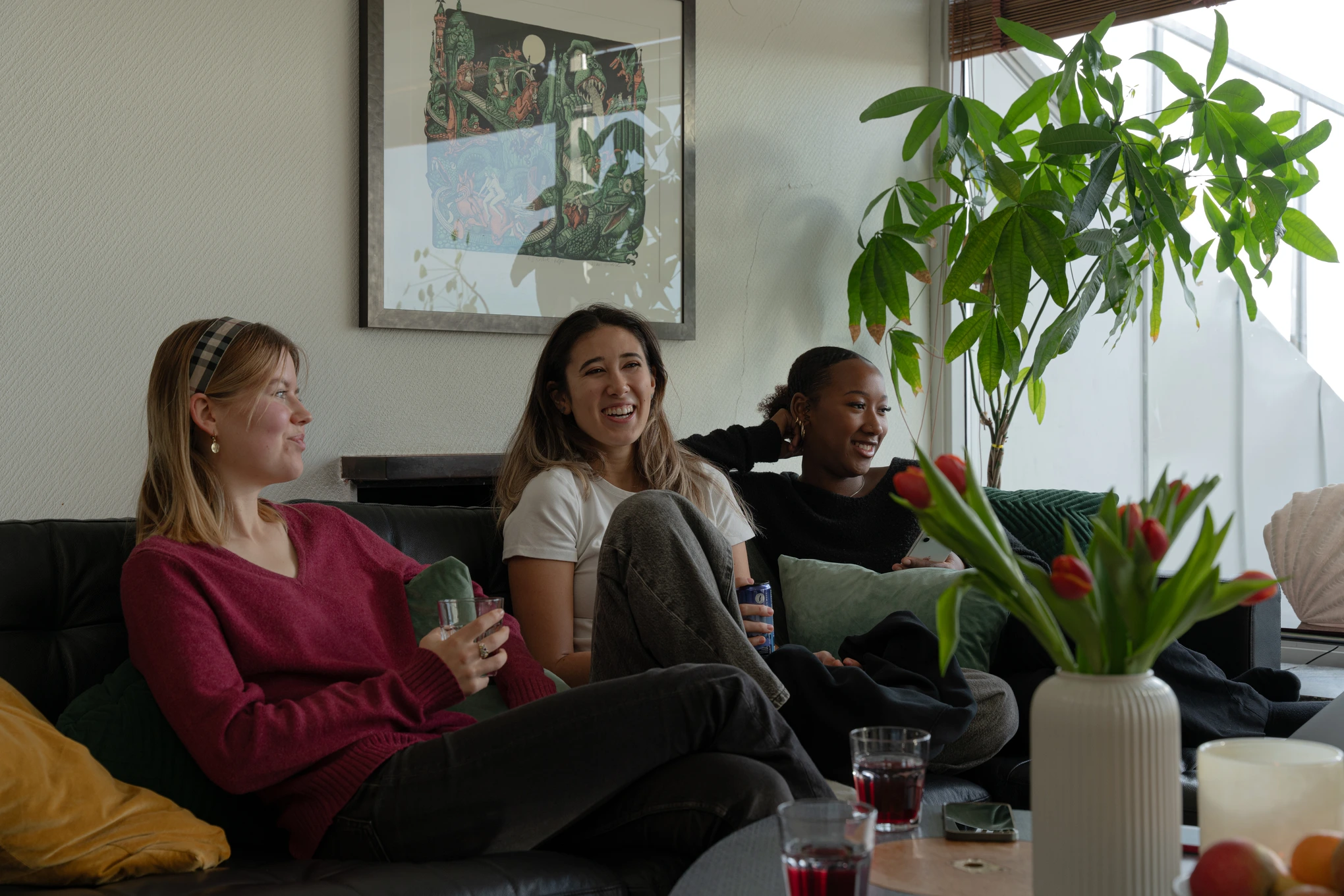 Tre personer sitter i en soffa i ett vardagsrum med gröna växter och tulpaner på bordet framför sig. De dricker och skrattar tillsammans i en avslappnad atmosfär.