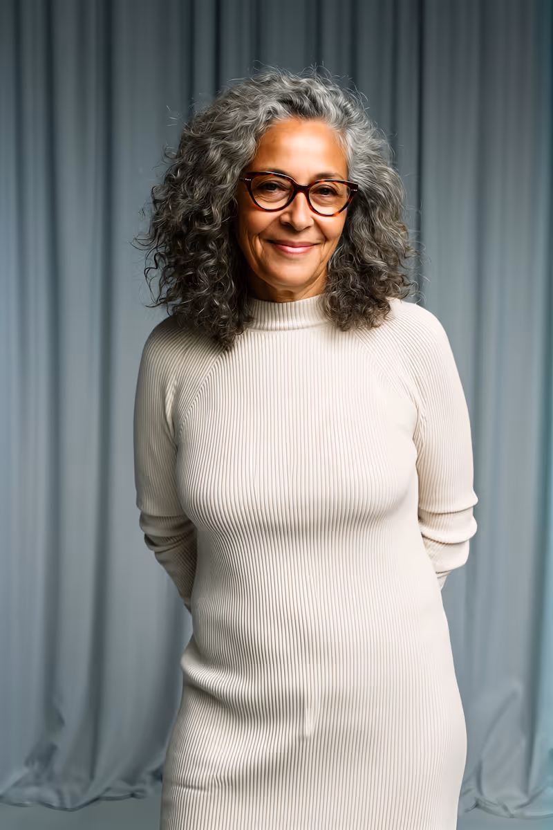 Smiling mature woman with curly gray hair wearing glasses and a cream ribbed dress standing against a blue-gray curtain background.