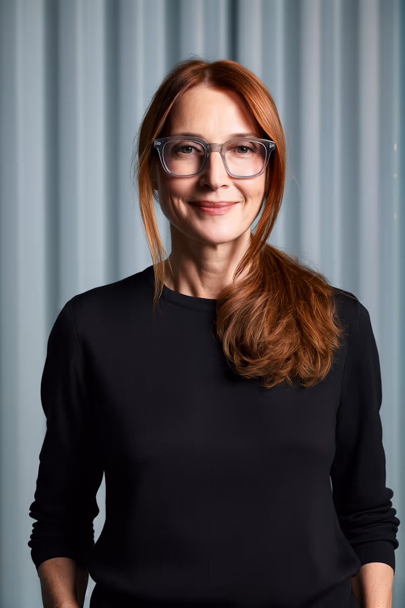 Smiling woman with long red hair tied to one side, wearing glasses and a black top.