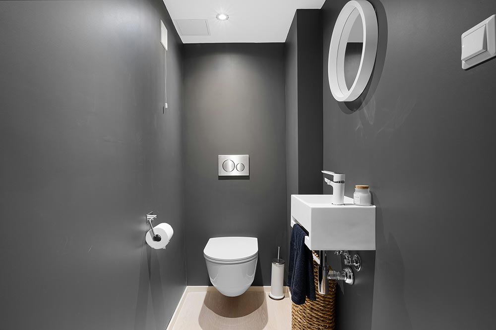 Minimalist bathroom remodel with a wall-mounted white toilet, small white sink, round mirror, and dark gray walls.