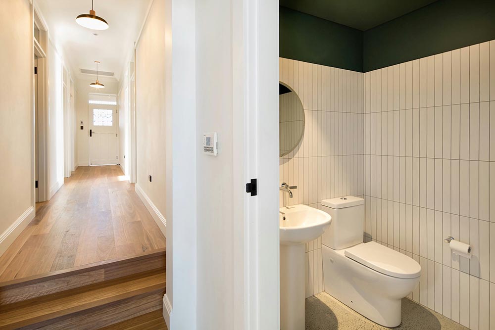 Bright hallway with wooden floors leading to a door and a small bathroom with white vertical tiles, a pedestal sink, and a toilet.