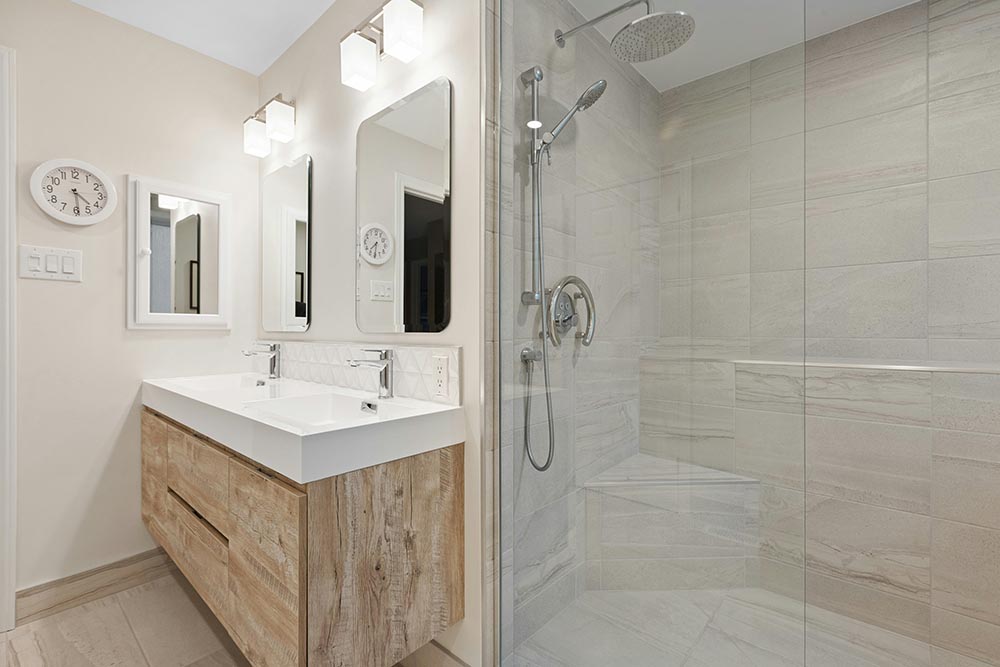 bathroom remodel with floating wooden vanity with double sinks and two rectangular mirrors, next to a glass-enclosed walk-in shower with rain and handheld showerheads.