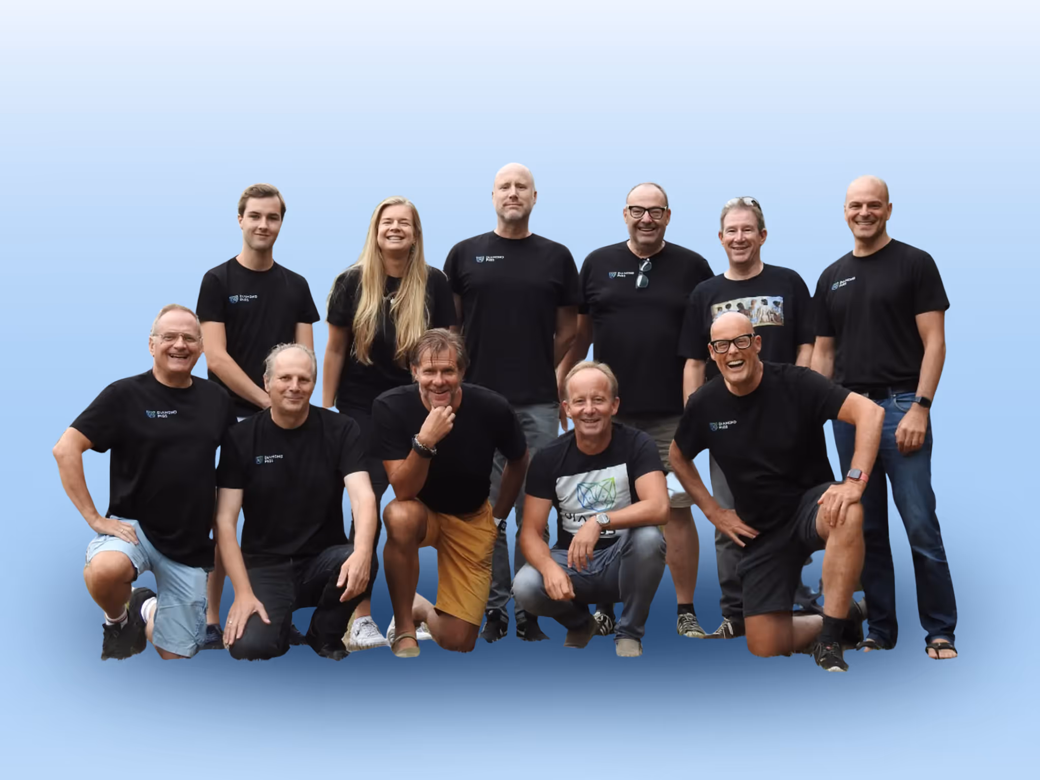Group of twelve smiling people posing together against a light blue background, wearing casual clothing, mostly black t-shirts.