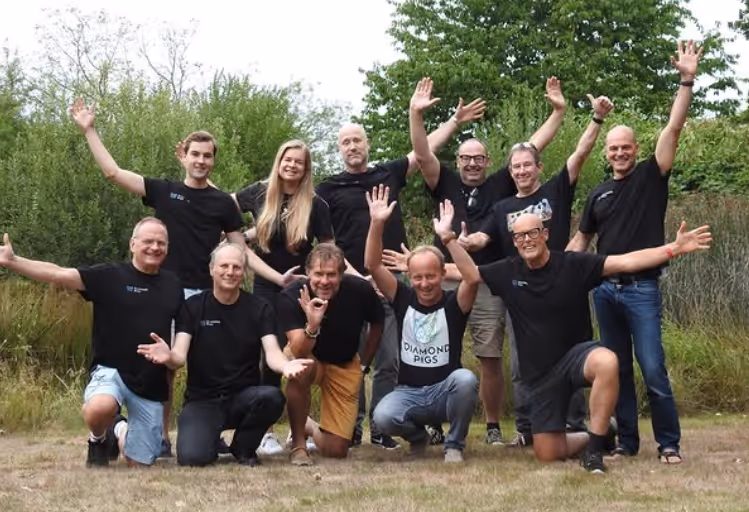 Group of twelve adults outdoors with trees in the background, smiling and raising their hands, some kneeling and some standing.