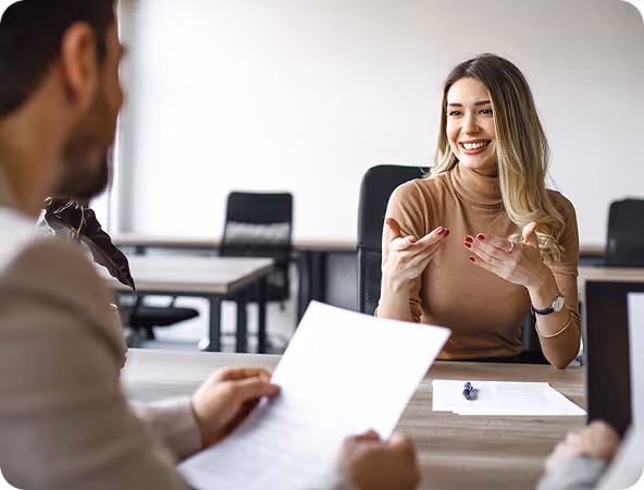Smiling woman in a brown turtleneck engaged in conversation with a man holding a document in an office setting.