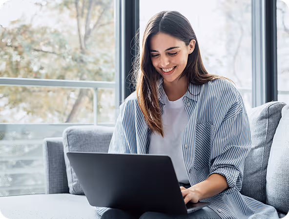 Smiling woman sitting on a couch using a laptop near large windows with a blurred outdoor background.