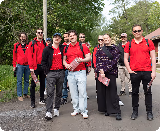 Group of young adults standing on a path outdoors surrounded by green trees, most wearing red shirts and holding pamphlets.