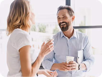 Two colleagues smiling and talking while holding coffee cups in an office setting.