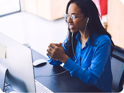 Smiling woman wearing glasses and earphones, sitting at a desk and looking at a computer screen.