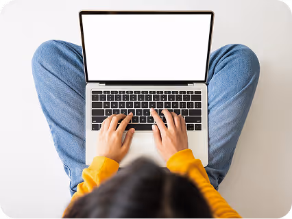 Person wearing a yellow sweater typing on a laptop while sitting cross-legged on the floor.