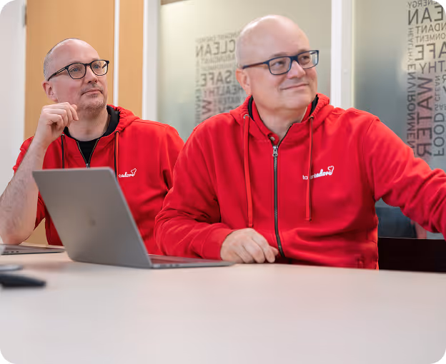 Two men in matching red hoodies sitting at a table with a laptop, looking attentively to the side in a modern office setting.