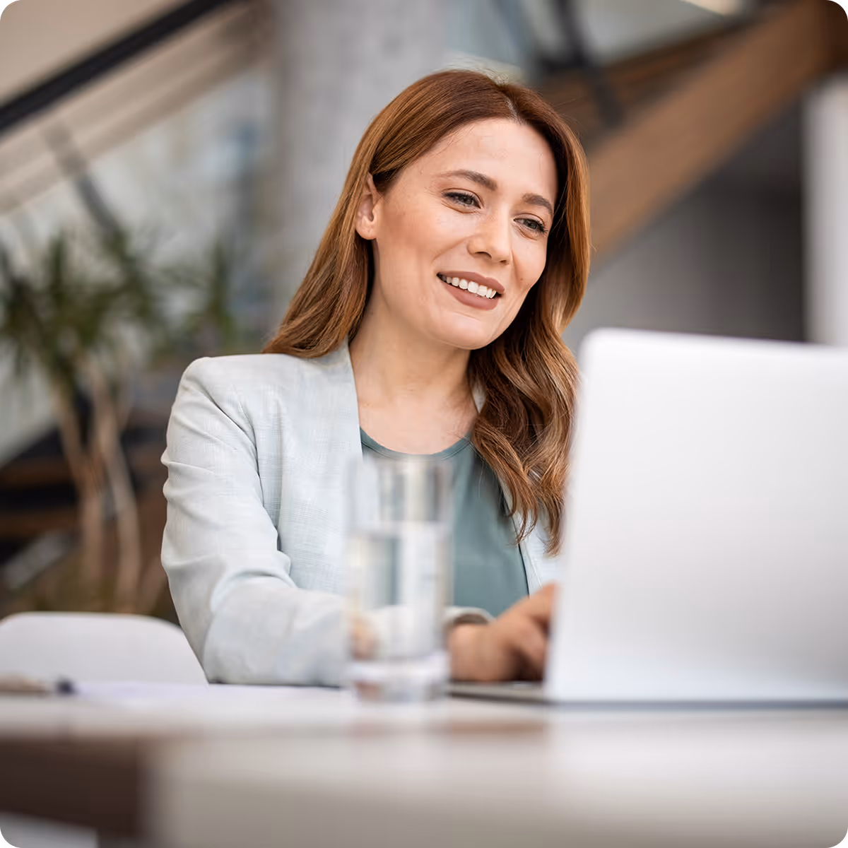 Smiling woman with red hair working on a laptop at a desk with a glass of water in an office.