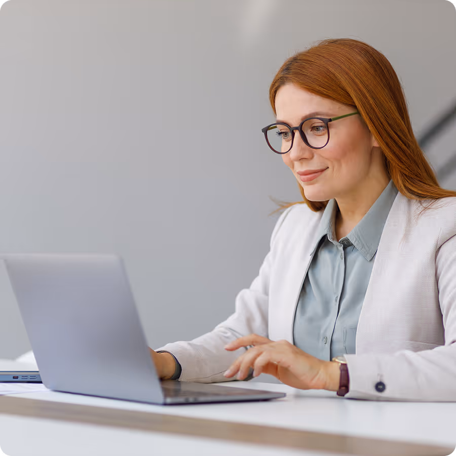 Smiling woman with red hair and glasses working on a laptop at a white desk.