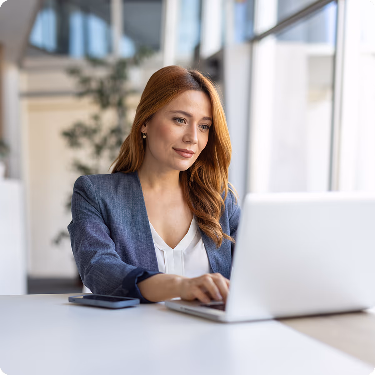 Woman with red hair in a blazer working on a laptop at a white desk next to a smartphone.