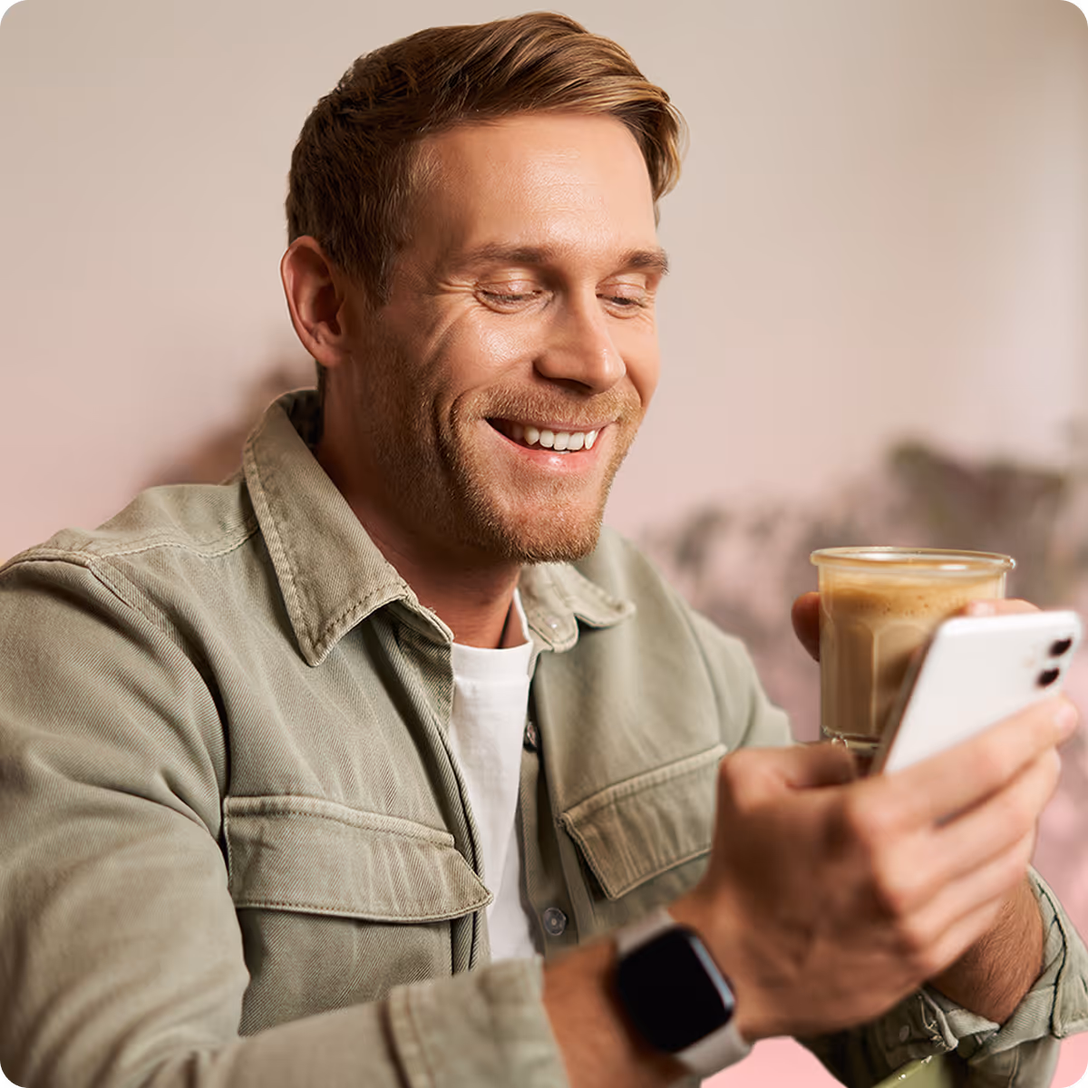 Smiling man with light brown hair wearing a green jacket, holding a small glass of iced coffee and looking at his smartphone.