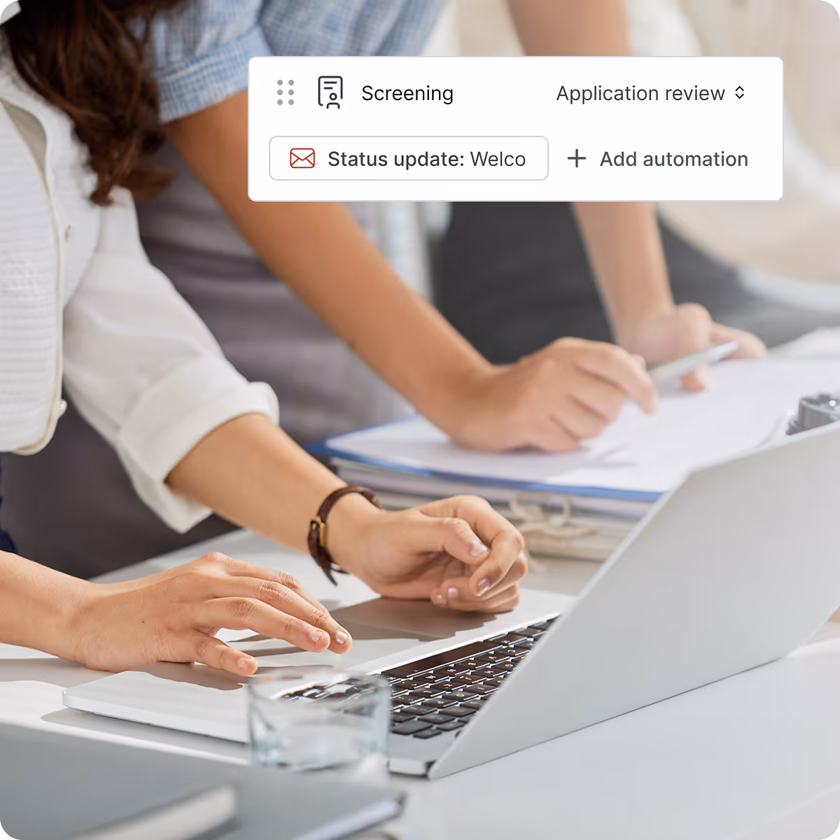Two people working together at a desk with one typing on a laptop and the other holding a pen over documents.