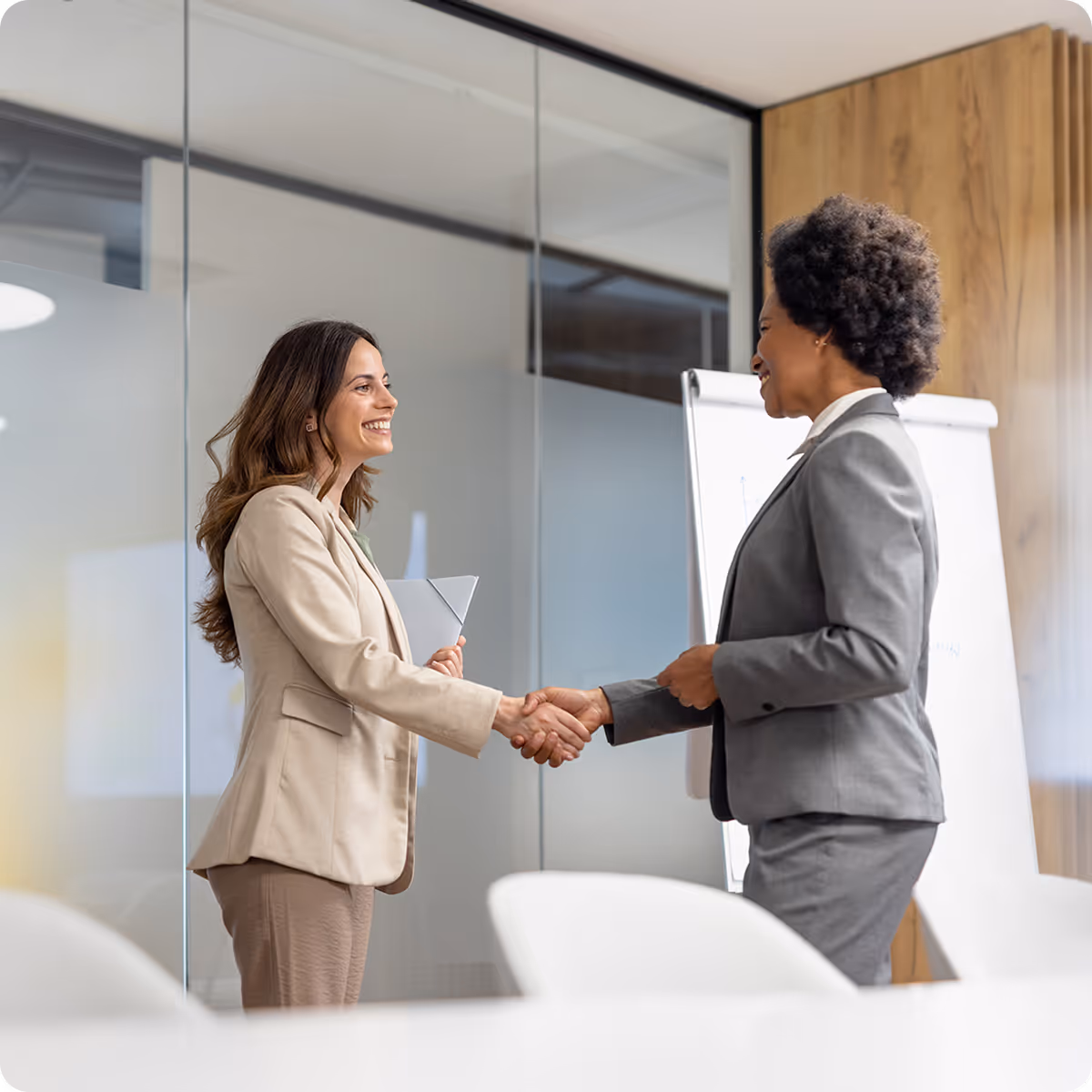 Two professional women in business attire shaking hands and smiling in a modern office.