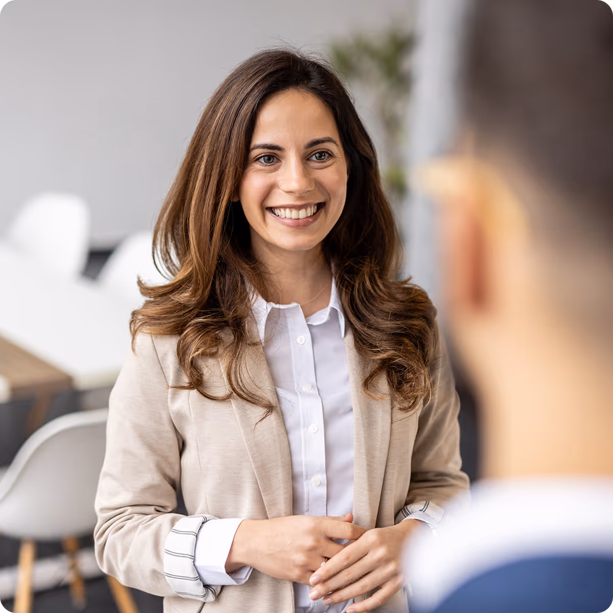 Smiling businesswoman with long brown hair, wearing a beige blazer and white shirt, engaged in conversation in an office.