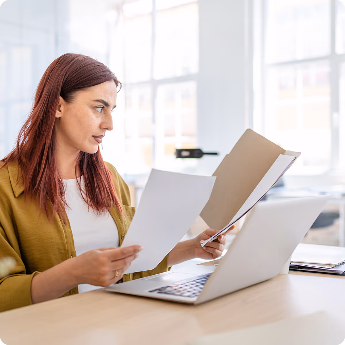 Woman with red hair reviewing documents at a desk with a laptop in a bright office.