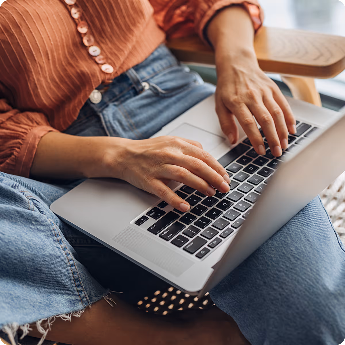 Person wearing a rust-colored shirt and blue jeans typing on a laptop keyboard in their lap.