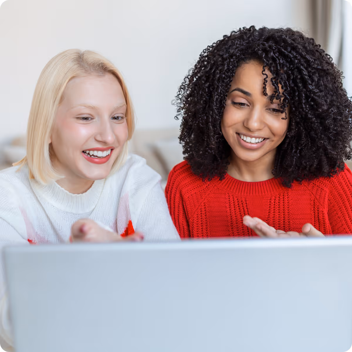 Two women smiling and looking at a laptop screen, one wearing a red sweater and the other a white sweater.