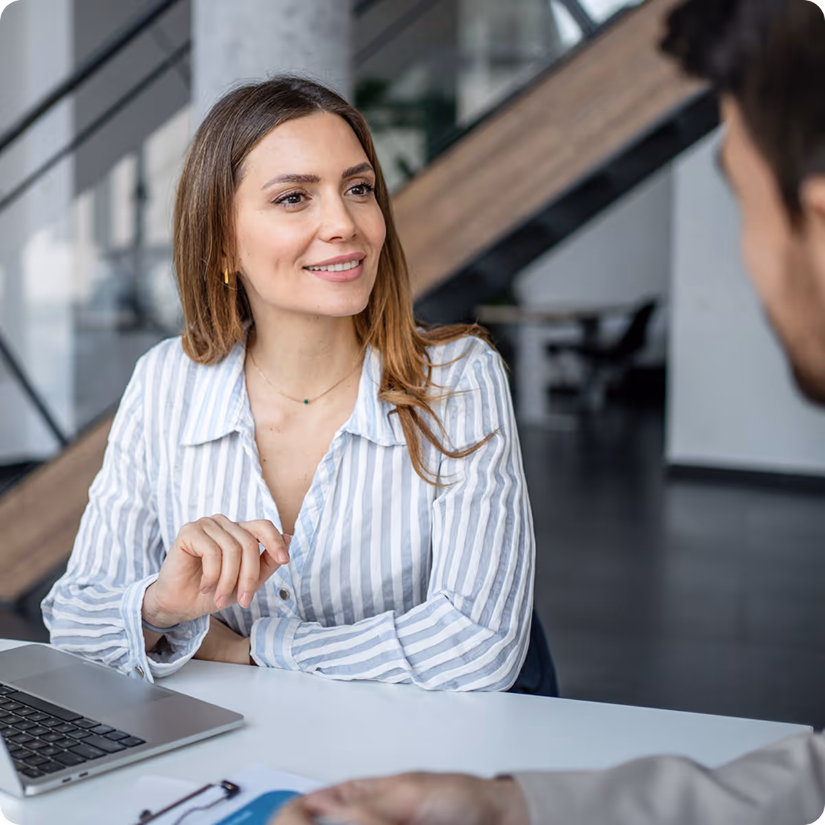 Smiling woman in a striped shirt sitting at a desk with a laptop, talking to a man in an office.