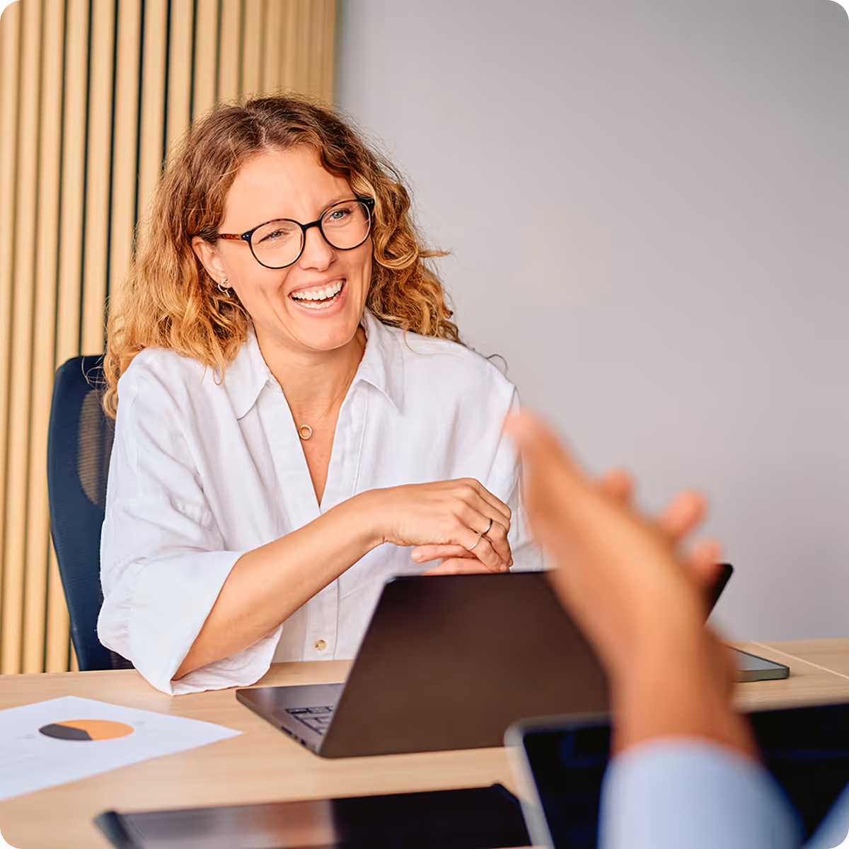 Smiling woman with glasses and curly hair sitting at a desk with a laptop, engaged in conversation.