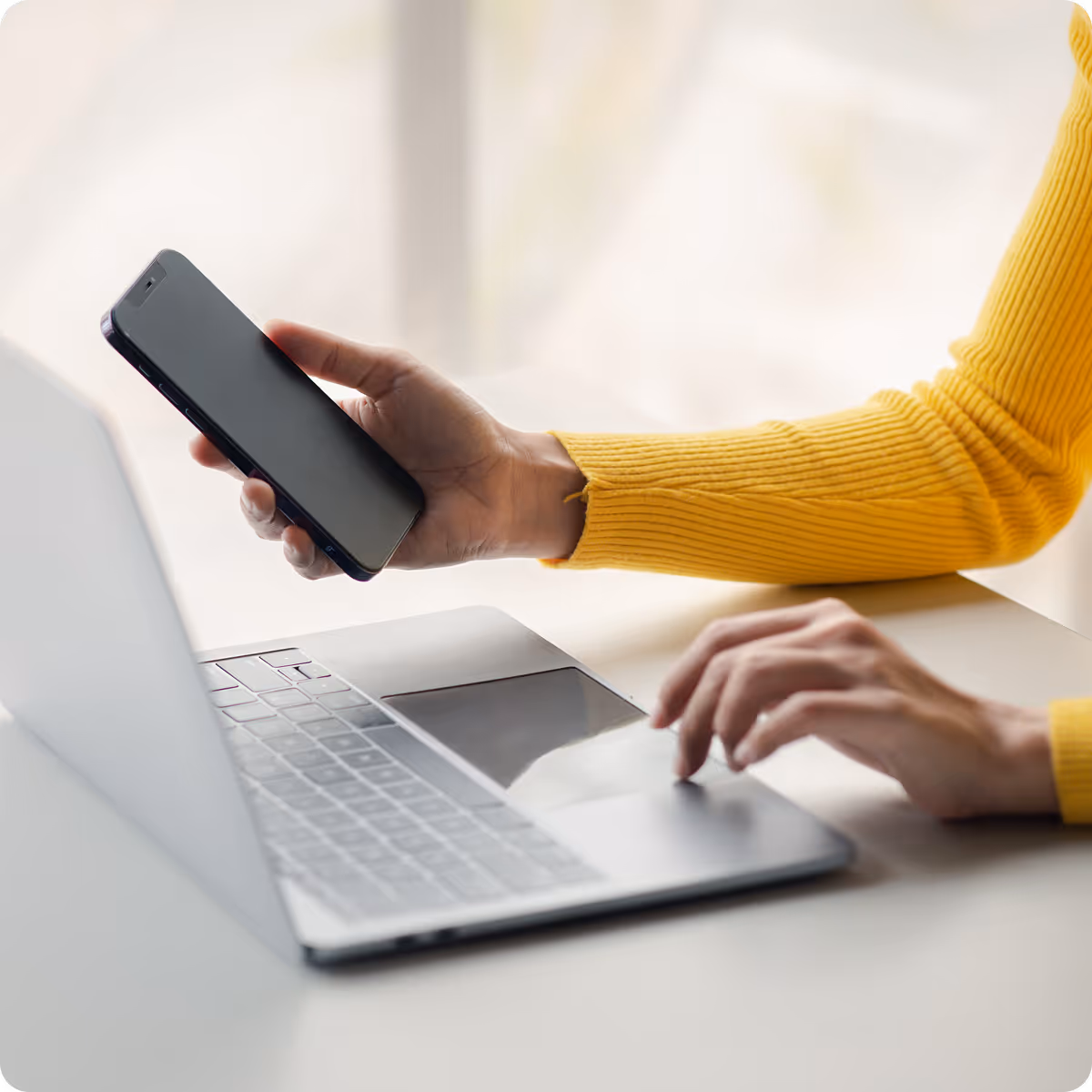 Person wearing a yellow sweater holding a smartphone in one hand and using a laptop with the other hand on a white desk.