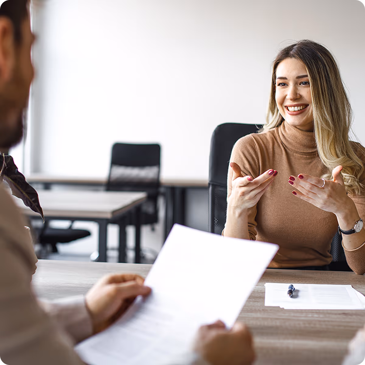 Smiling woman with long hair and brown turtleneck engaging in conversation during a job interview with a man holding documents.