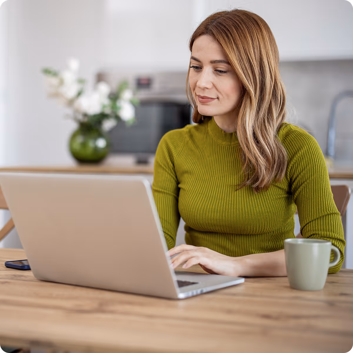 Woman with light brown hair wearing a green ribbed sweater working on a laptop at a wooden table with a green mug nearby.