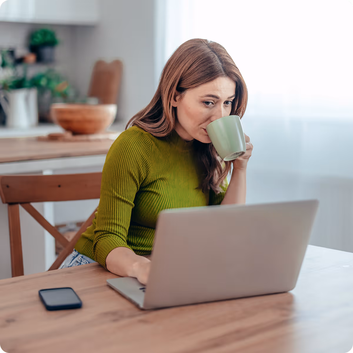Woman in green sweater drinking from a mug while working on a laptop at a wooden table.