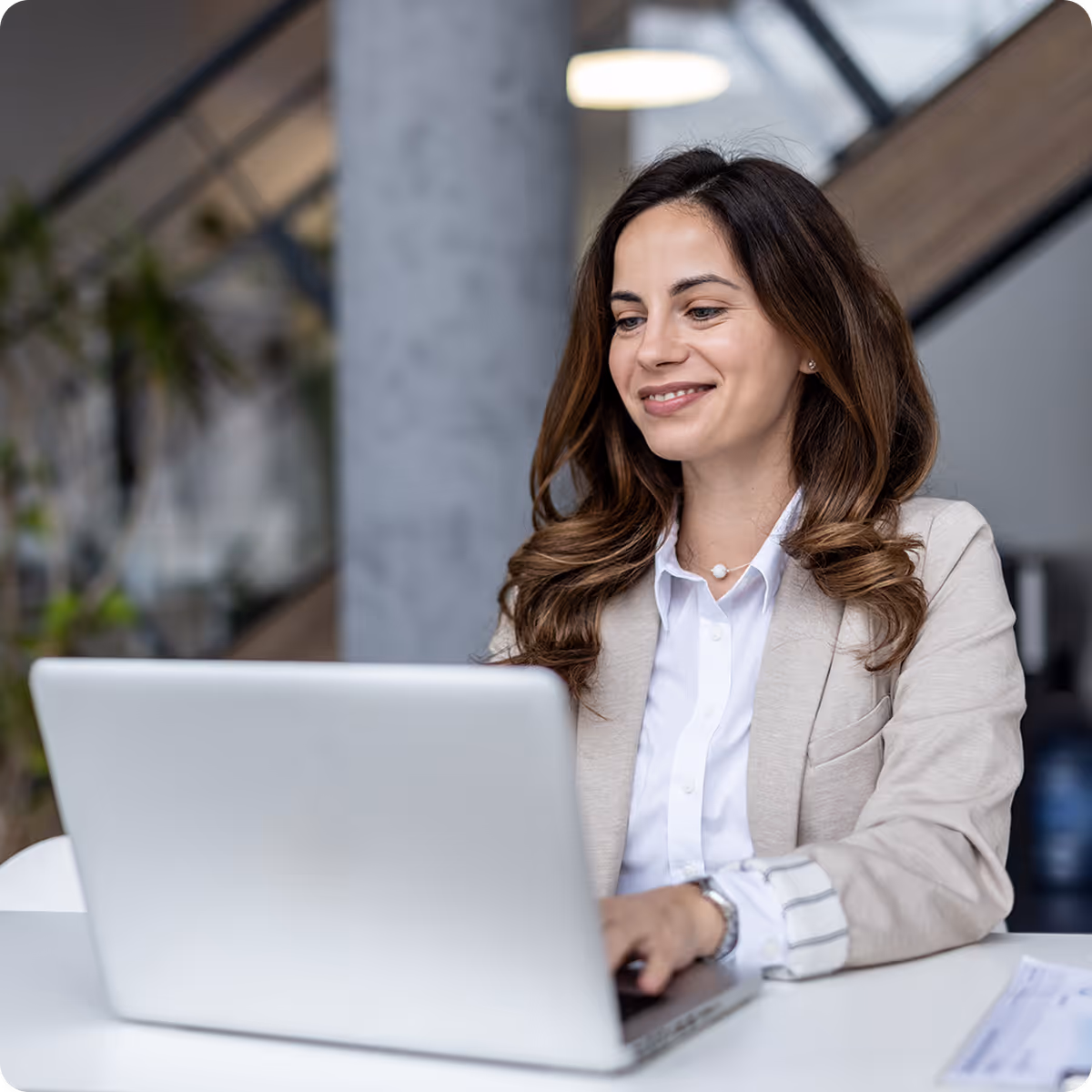 Smiling businesswoman in a beige blazer working on a laptop at a white desk in a modern office.