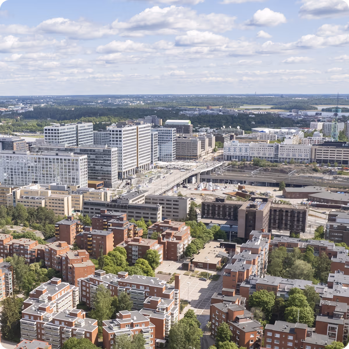 Aerial view of a cityscape with modern white office buildings, red residential buildings, green trees, and a partly cloudy sky.