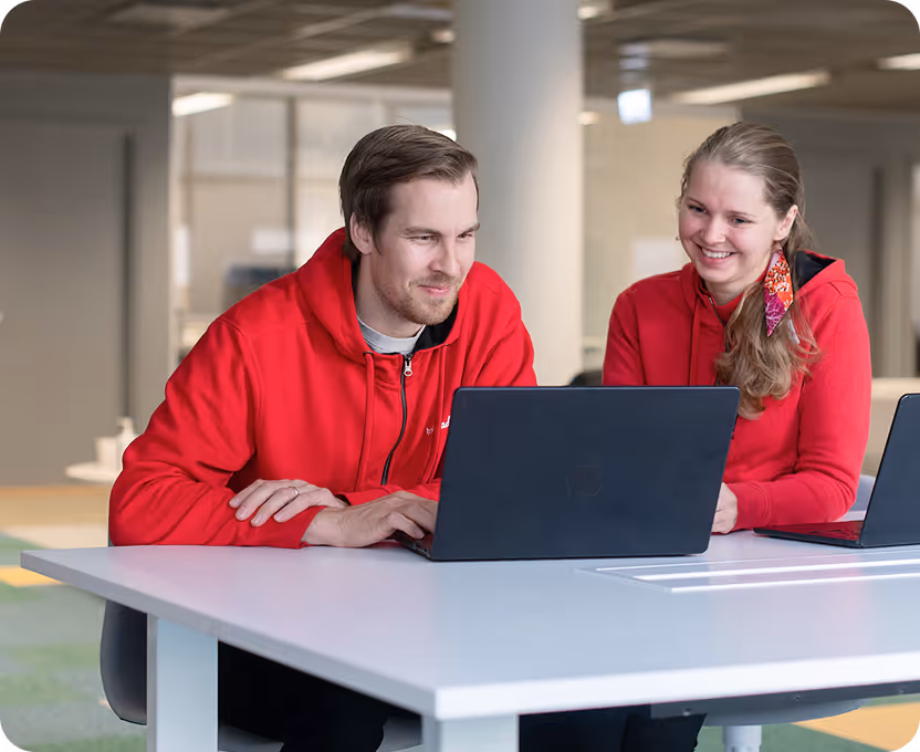A man and woman in red hoodies smiling and working together on a laptop at a white table in a modern office.