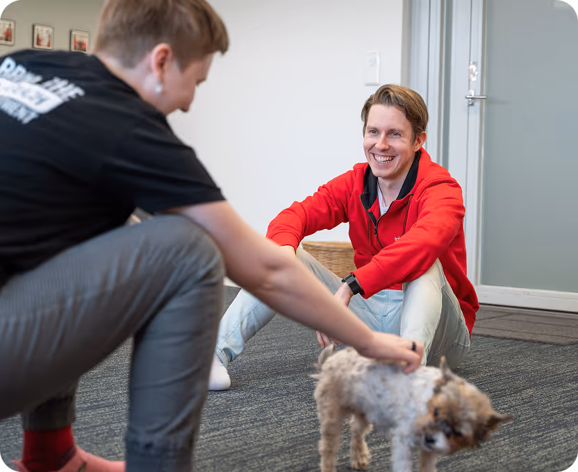Two people sitting on a carpeted floor petting a small dog, one wearing a red jacket and smiling.