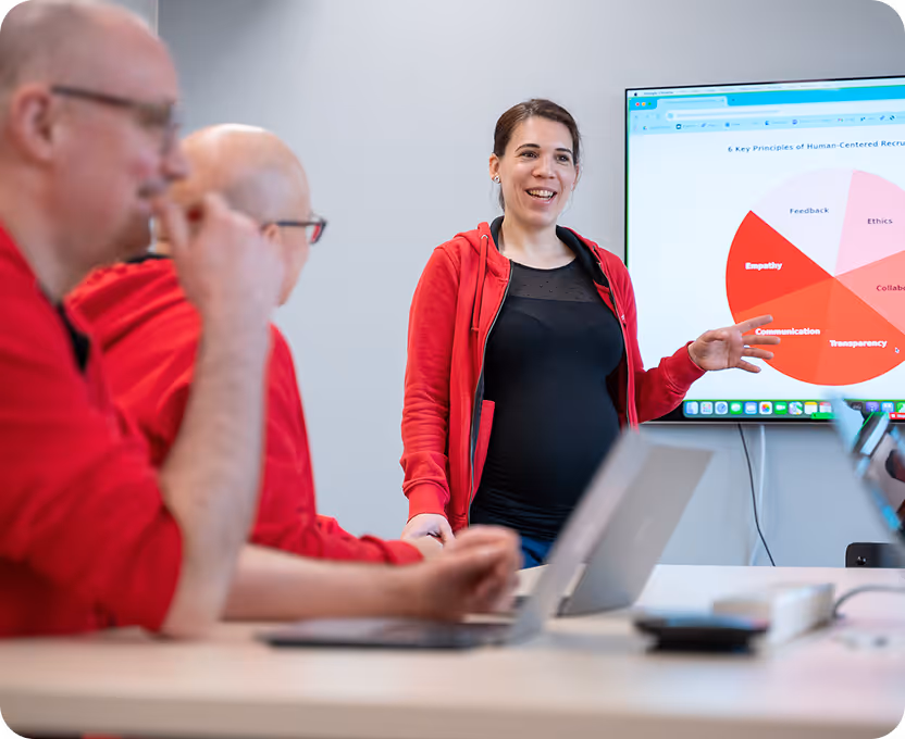 Woman in a red jacket presenting a pie chart about human-centered recruitment to a group of men in red shirts.