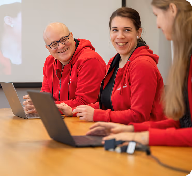 Three people in red hoodies sitting at a table with laptops, smiling and engaging in a discussion.