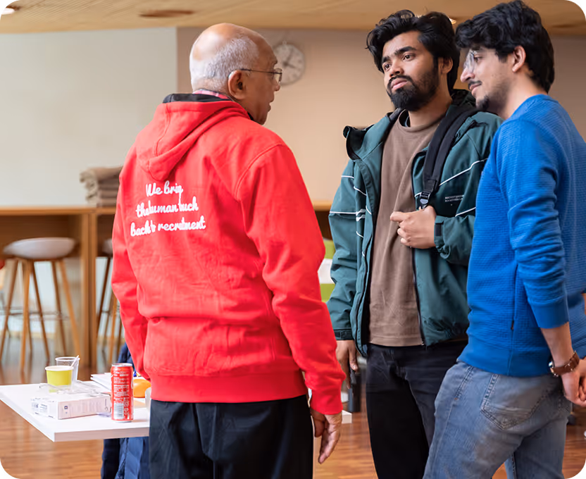 Three men standing and talking indoors, one wearing a red hoodie with partially visible white text on the back, and two others dressed casually with backpacks.