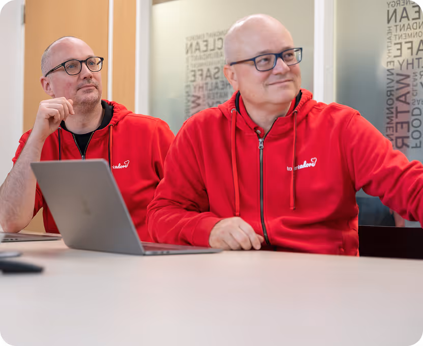 Two men wearing red hoodies with logos sitting at a table with a laptop in a meeting room.