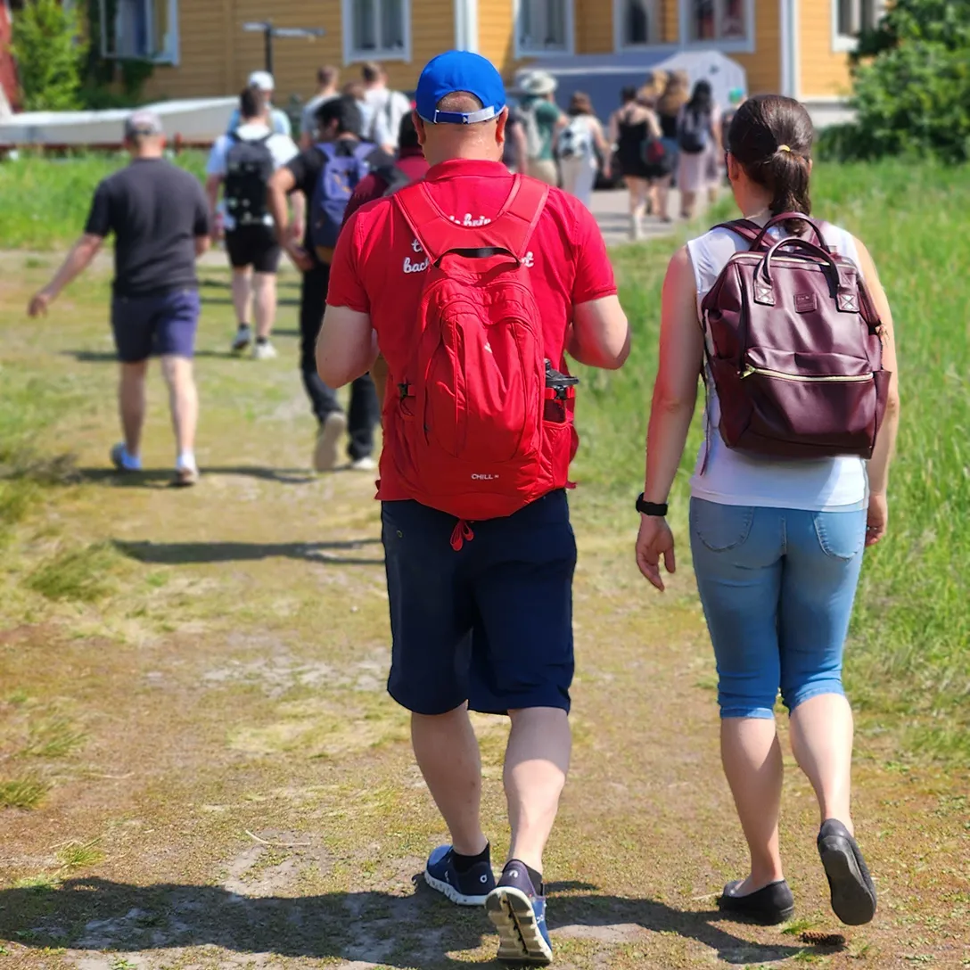 Group of people walking outdoors on a sunny day on a path with grass and a yellow building in the background.
