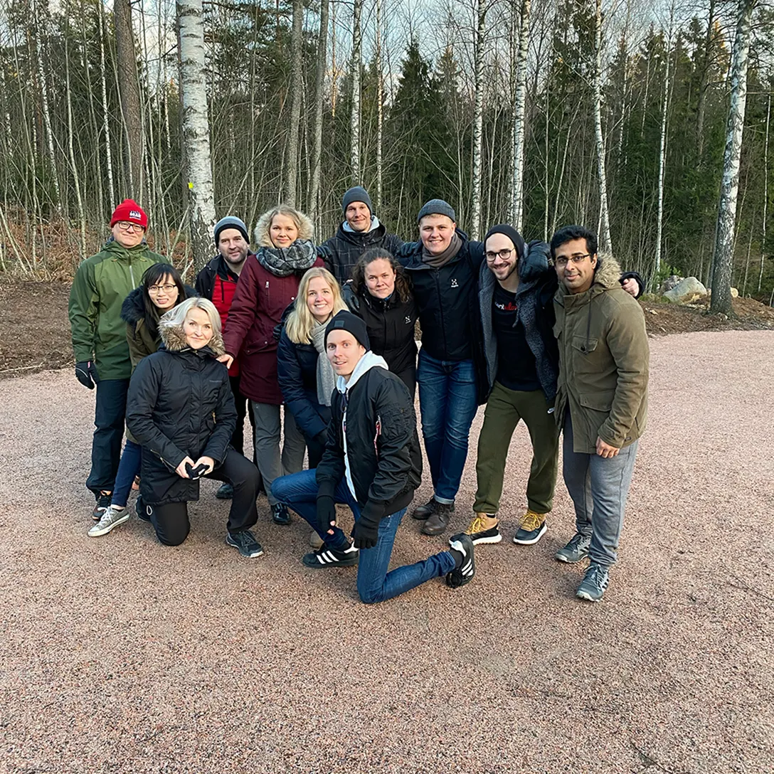 Group of twelve adults dressed in winter clothing posing together outdoors on a gravel path with leafless birch trees in the background.