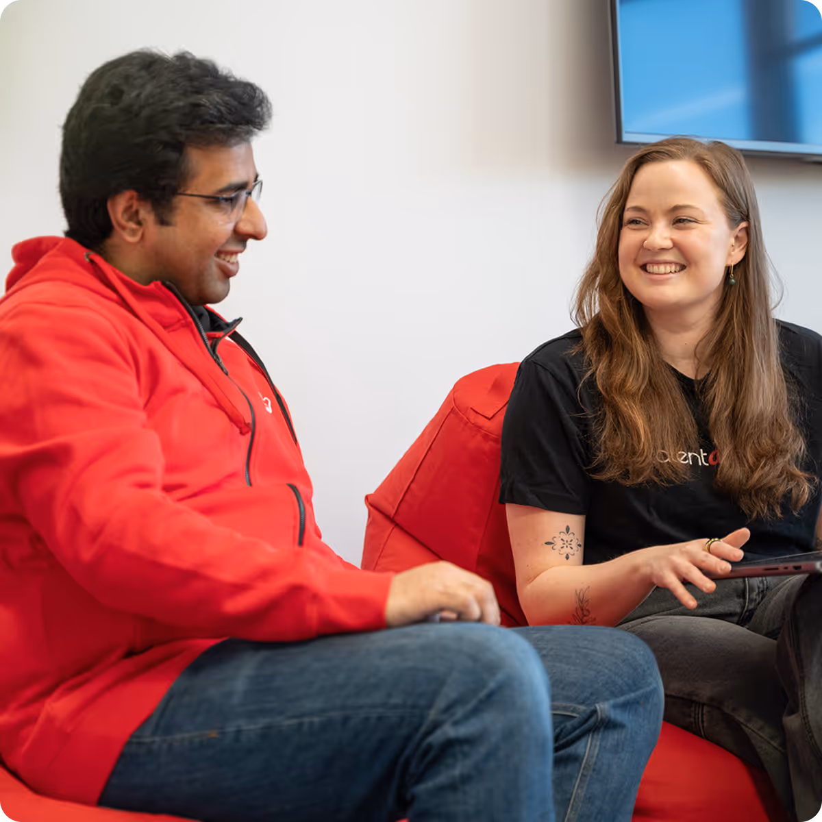 A man in a red hoodie and glasses smiling and talking to a woman with long brown hair, wearing a black t-shirt, sitting on red chairs.