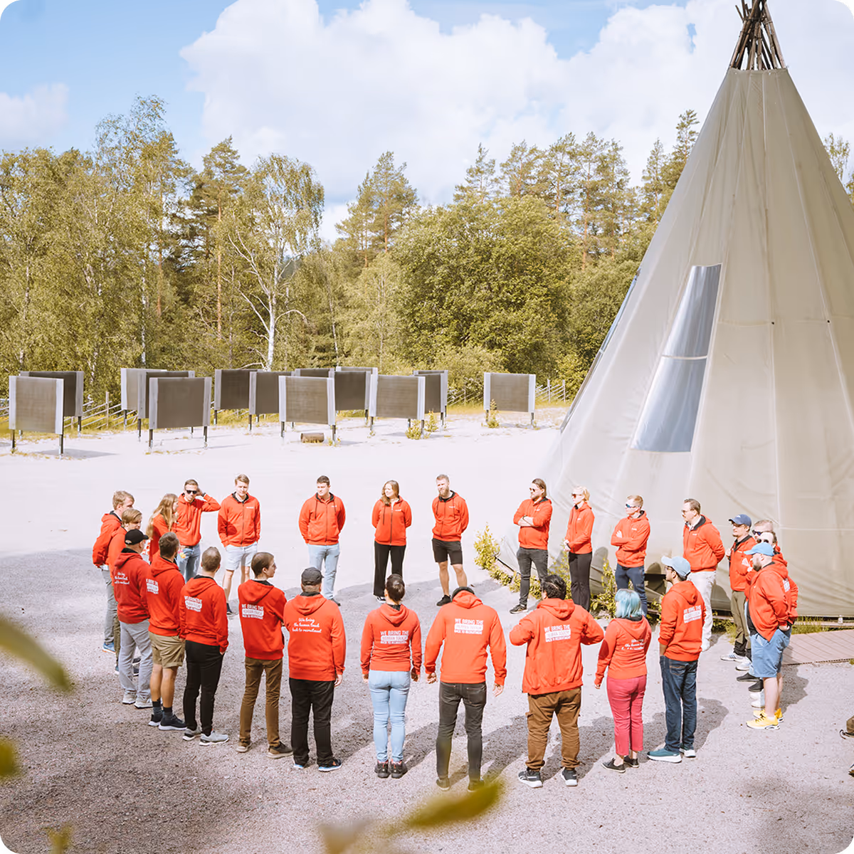 Group of people standing in a circle outdoors near a large teepee, all wearing matching red hoodies in a wooded area.