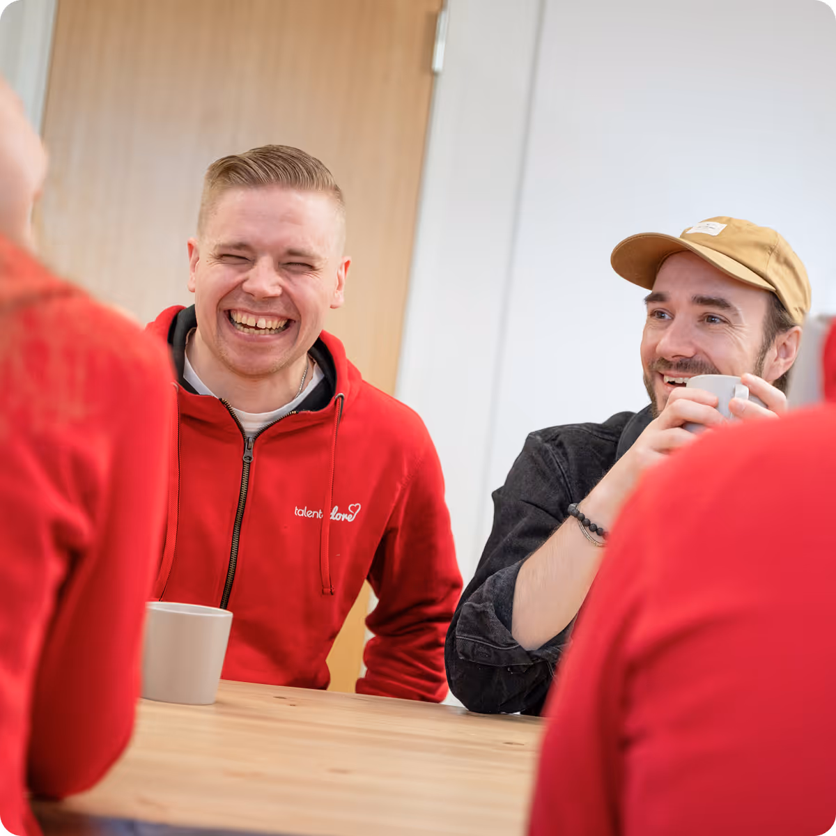 Two men sitting at a wooden table laughing and holding mugs, one wearing a red hoodie and the other a beige cap and black shirt.