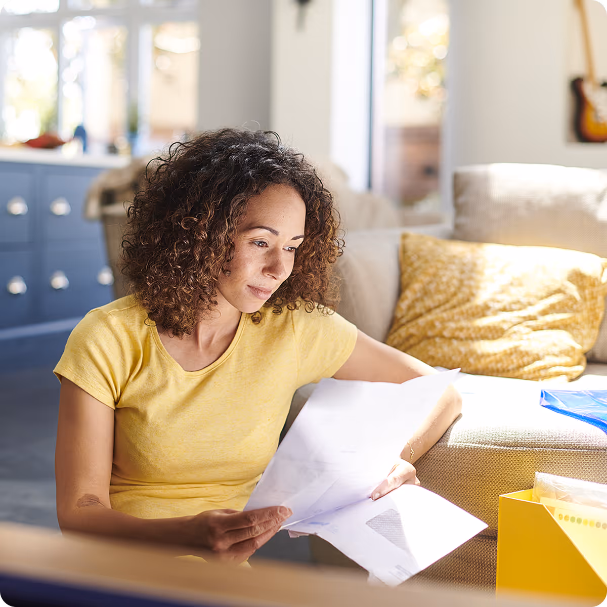 Woman in a yellow shirt sitting on the floor near a couch, reading papers in a bright living room.